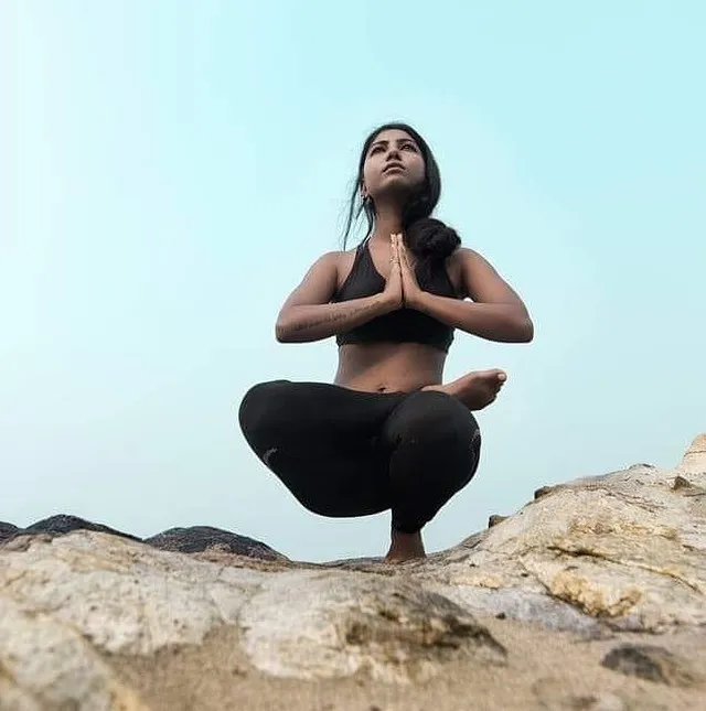 Afsha in a low squat with hands at the chest, balanced on rock against an open sky.
