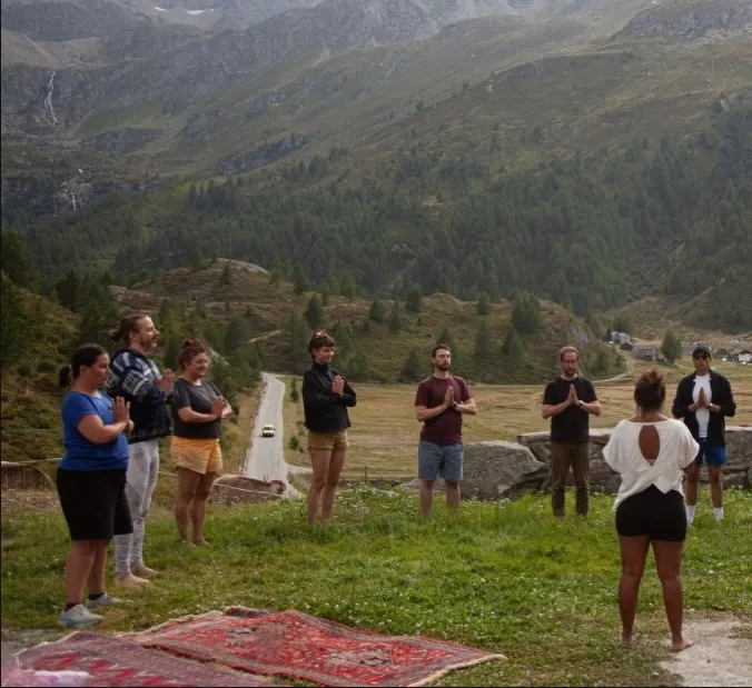 A group standing in a circle at the start of an outdoor class, hands at the chest.